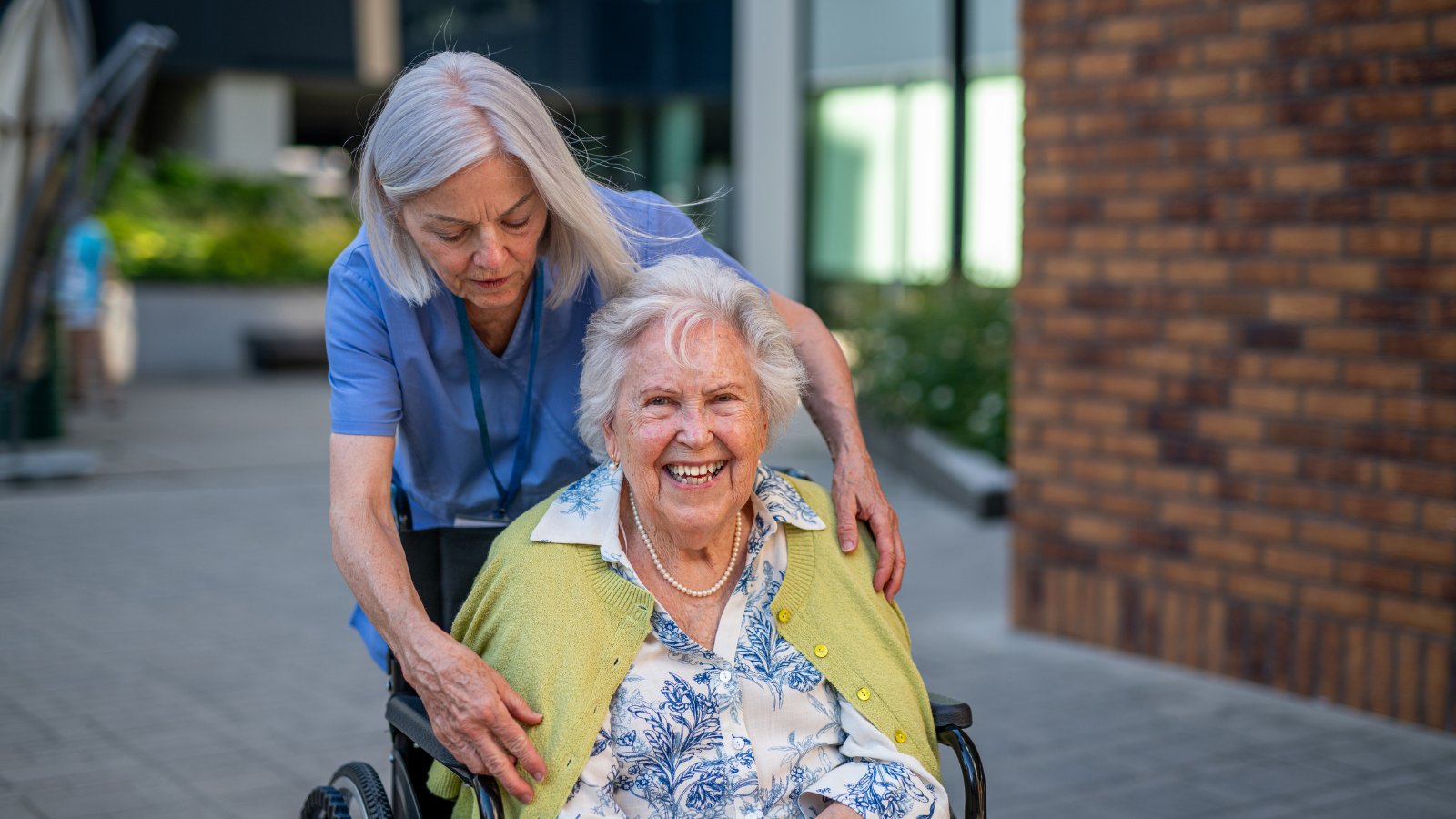 Caregiver assisting smiling elderly woman in wheelchair outside medical facility